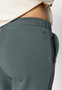 Tumši pelēki treniņbikšu izgatavoti no mīkstas auduma, ar auklu jostasvietā un sānu kabatām. Vienkāršs dizains ar mazu logotipa detaļu.