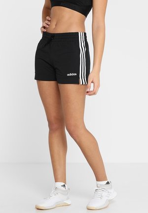 Kvinde iført sorte Adidas-shorts med hvide striber og logo, kombineret med hvide Adidas-sneakers og strømper, stående mod en grå baggrund.