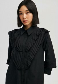 Svart skjorta med strukturerade, diagonala veck på framsidan och ärmarna, med krage och knäppning. Slät tygtextur.