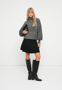 Pull en maille texturée gris, jupe évasée rayée noir et blanc, bottes noires à talons arrivant au genou, tenant un sac à main noir.