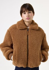 Chaqueta de forro polar marrón con cuello, cierre de cremallera y puños acanalados. Es de corte oversized con una superficie texturizada y esponjosa para mayor calidez.