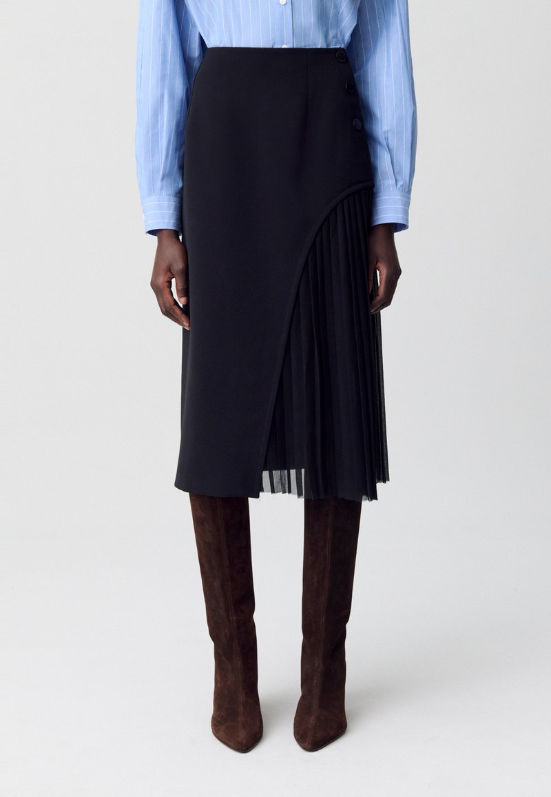 Gonna nera con sovrapposizione trasparente plissè, dotata di bottoni laterali e una silhouette elegante; abbinata a una camicia a righe blu chiaro e stivali marroni al ginocchio.