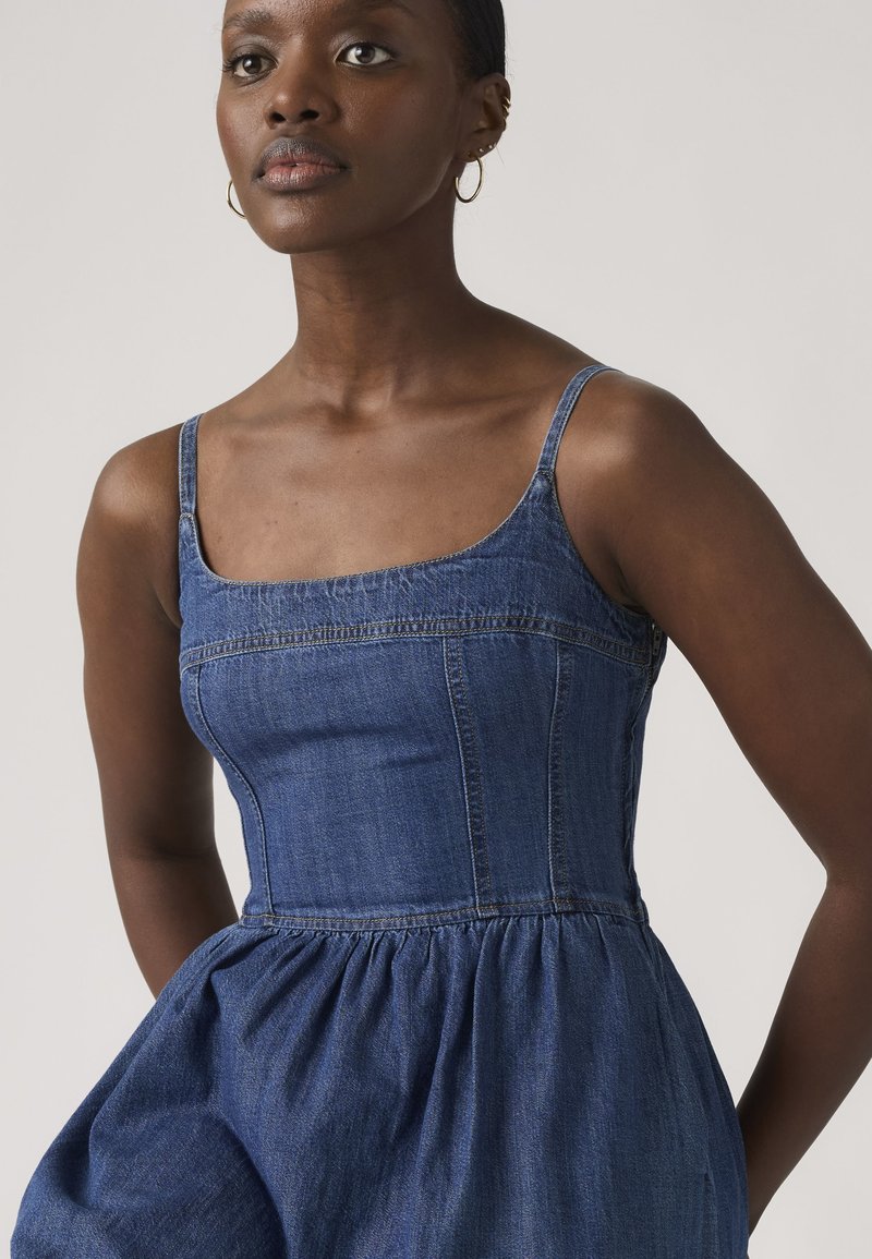 Woman with dark skin wearing sleeveless blue denim dress with fitted bodice and flared skirt, standing against light background.