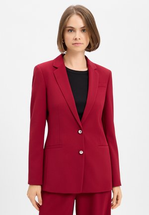 Vrouw met kort bruin haar draagt een aansluitend rood colbert en bijpassende broek over een zwarte top op een effen lichte achtergrond.