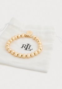 Pulseira de contas de ouro repousando sobre um saquinho branco com o logótipo "RLL". Superfície suave e brilhante com um fecho de bico para um fechamento seguro.