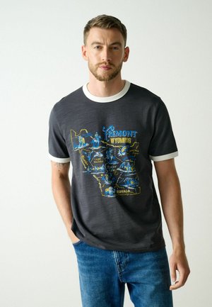 Mand iført mørkegrå T-shirt med hvid kant og blå-gul Fremont Wyoming kortdesign, kombineret med blå jeans, står mod en hvid baggrund.