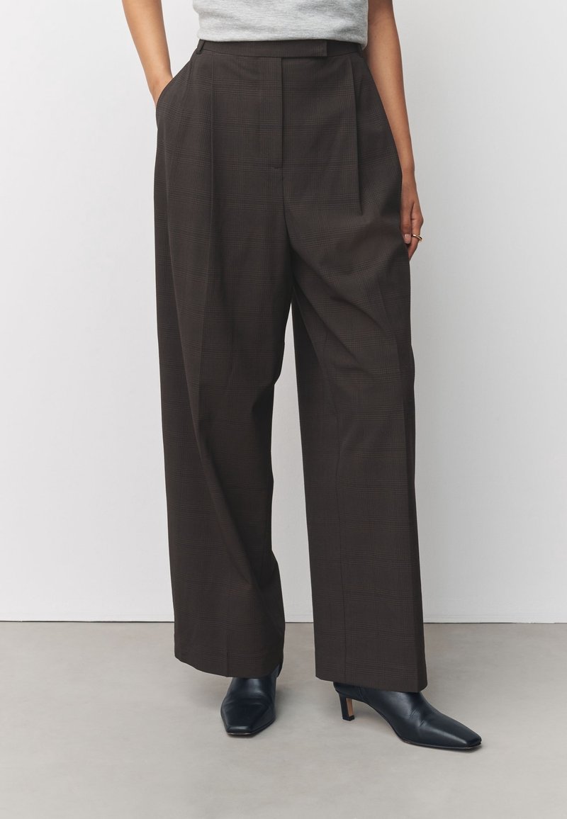 Femme portant un pantalon à carreaux foncé taille haute et jambes larges, un top gris rentré, et des bottines à talons en cuir noir pointues.