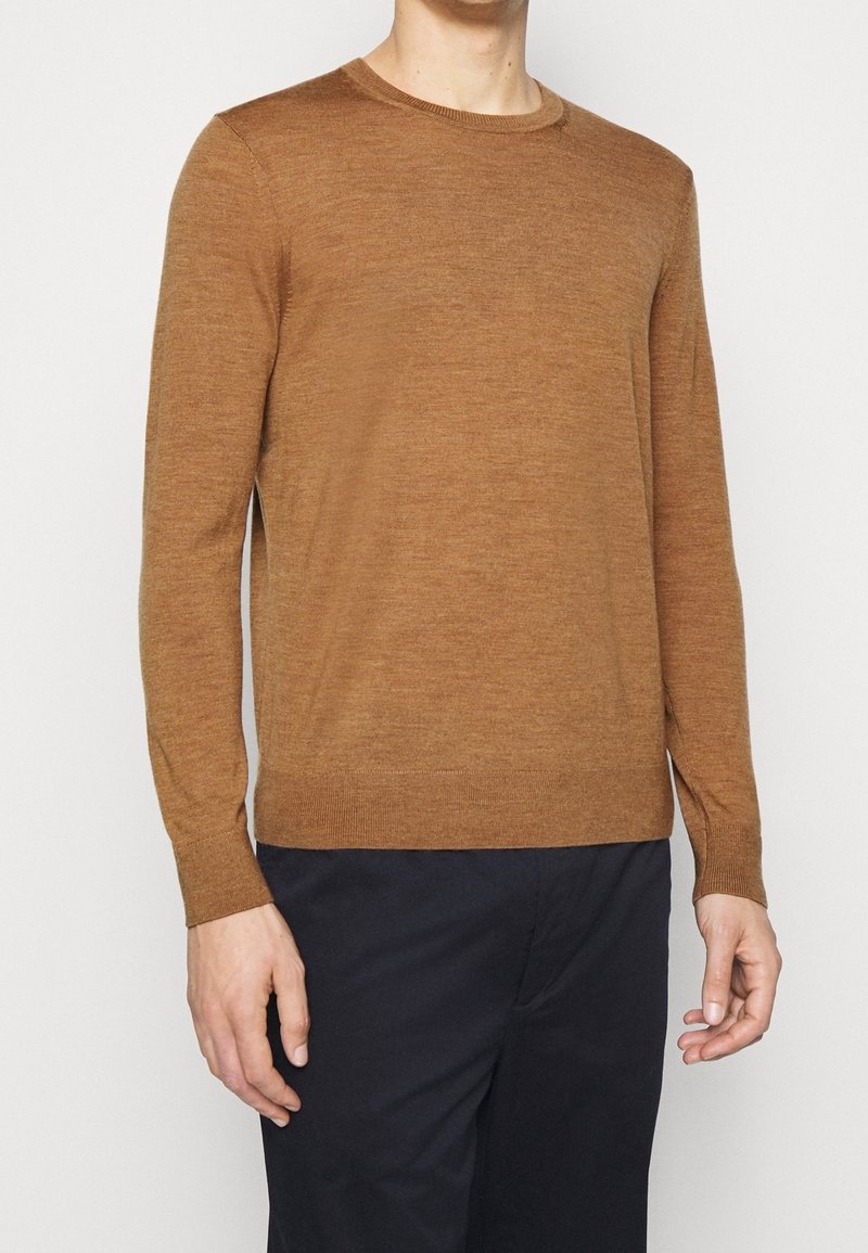 Homme portant un pull à manches longues marron uni et un pantalon noir, debout devant un fond clair uni.