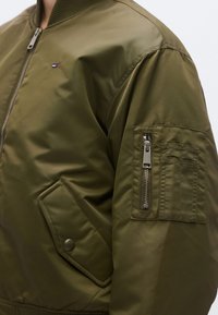 Χακί πράσινο bomber μπουφάν με κλείσιμο με φερμουάρ, μανσέτες με ρίγες και τσέπη μανικιού με φερμουάρ και ορθογώνιο καπάκι τσέπης.