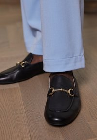 Schwarze Lederloafers mit goldener Pferdebitte, kombiniert mit hellblauen Hosen, auf einem Holzfußboden.