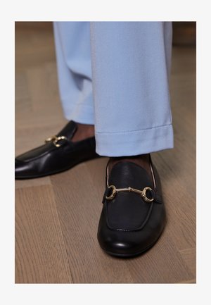 Schwarze Lederloafers mit goldener Pferdebitte, kombiniert mit hellblauen Hosen, auf einem Holzfußboden.