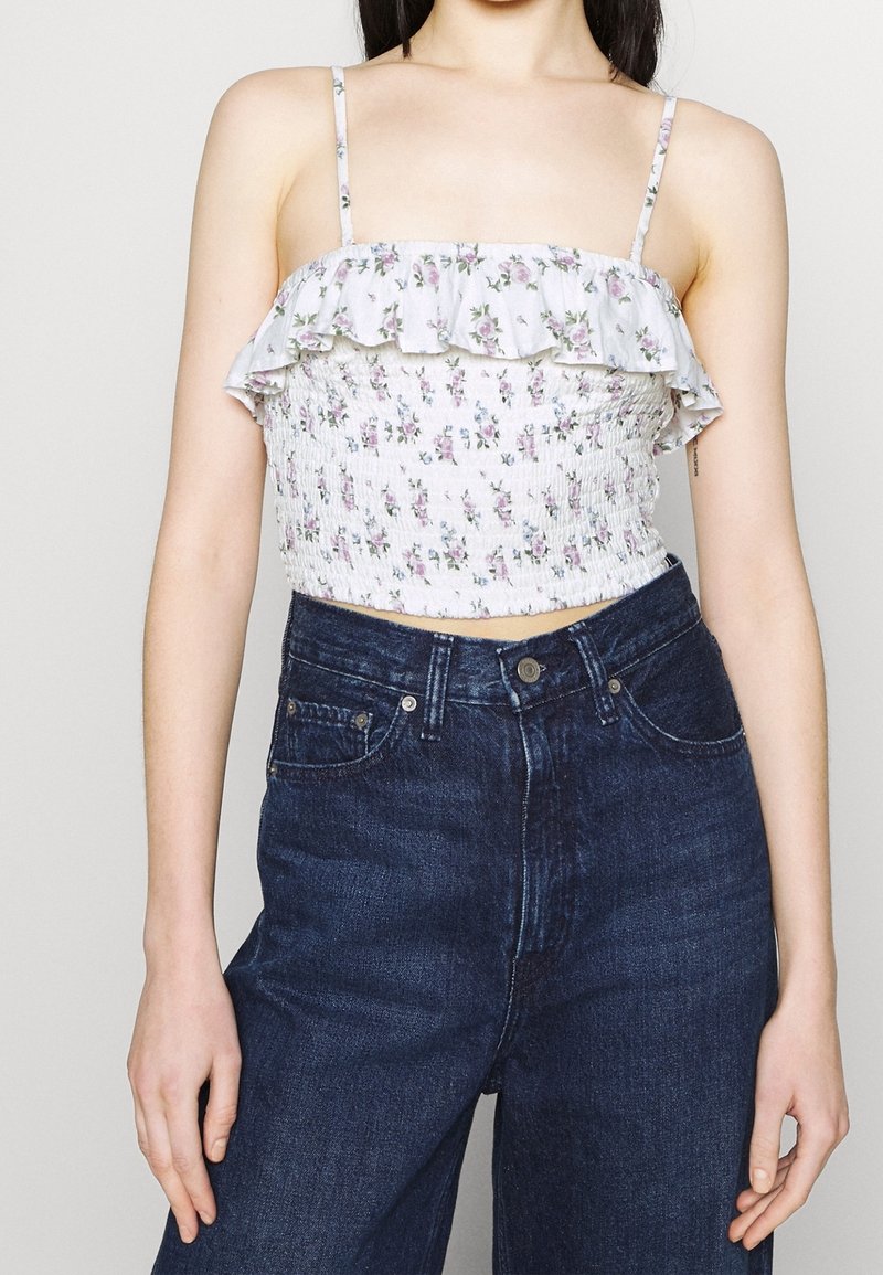 Květinový crop top s volánkovým výstřihem, elastickým korzetem a tenkými ramínky; zdobený malými fialovými a zelenými květinovými vzory na světlém pozadí.