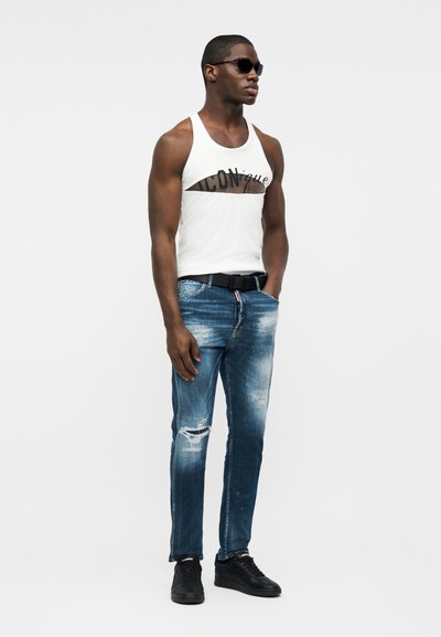 Άνδρας που φοράει λευκό αμάνικο μπλουζάκι με το κείμενο «ICON», φθαρμένο μπλε τζιν, μαύρη ζώνη, μαύρα αθλητικά παπούτσια και γυαλιά ηλίου, στέκεται με τα χέρια στις τσέπες.