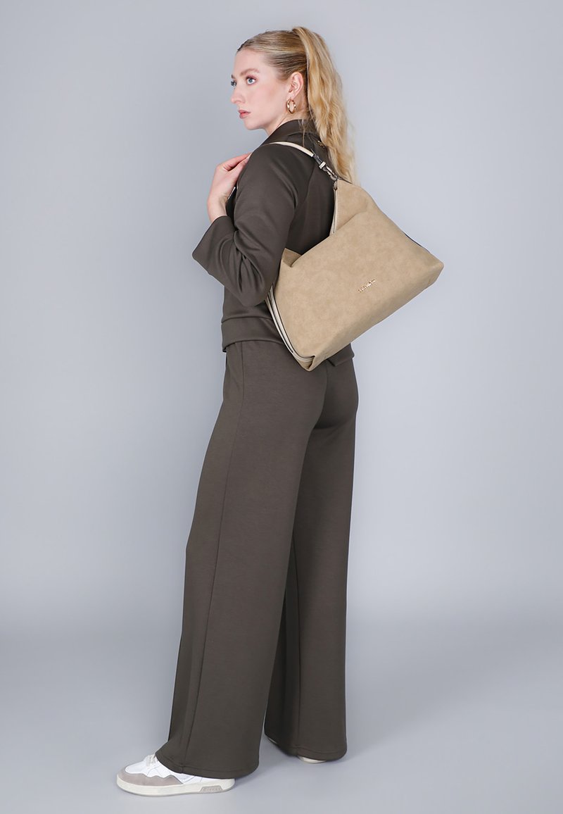 Beżowa torebka z zamszowym wykończeniem, z detalem w kolorze srebrnym przy zamku błyskawicznym oraz złotym logo. Nosi na pasku na ramię. Model w ciemnozielonym stroju.