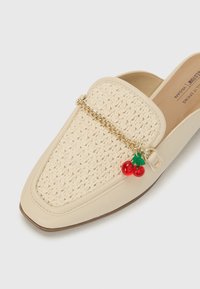 Zapato beige de tipo slip-on con una parte superior de rafia tejida, acento de cadena dorada y un colgante de cereza en rojo y verde. Diseño de puntera cuadrada.