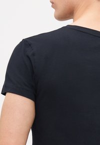 Hombre de perfil con una camiseta negra lisa de manga corta visible desde el hombro hasta la mitad de la espalda.
