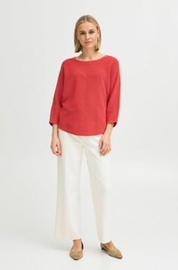 Pull en tricot rouge à la coupe décontractée, avec un col rond large et des manches trois-quarts, assorti à un pantalon blanc à jambes larges et des chaussures beiges.