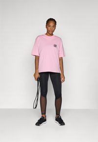 Tricou roz lejer din bumbac, cu un design grafic negru pe buzunar, asortat cu colanți negri cu accente din plasă și pantofi sport negri.