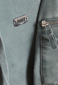 Lichtblauwe stof met een metalen label van het merk "JUICY" en een ritszak met een stoffen trekker.
