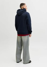 Tume sinine kapuutsiga jakk, millel on nöörid ja soonikkätised, punase särgi kohal, koos heleda laia jalgadega teksapükstega ja tumedate kingadega.