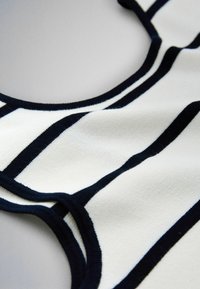 Material tricotat alb cu dungi negre îndrăznețe, orizontale și verticale, evidențiind o margine conturată și o textură moale pe o suprafață plată.