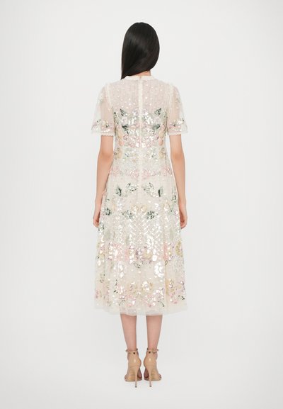 Beige jurk met bloemenborduursels en paillettenaccenten. Transparante korte mouwen, midi-lengte en golvende zoom. Draagt het met nude hoge hakken.