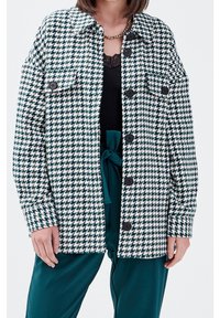 Groen-wit jasje met hondentandpatroon, knoopsluiting, twee voorzakken en een losse pasvorm. Draagt over een zwarte top en turquoise broek.