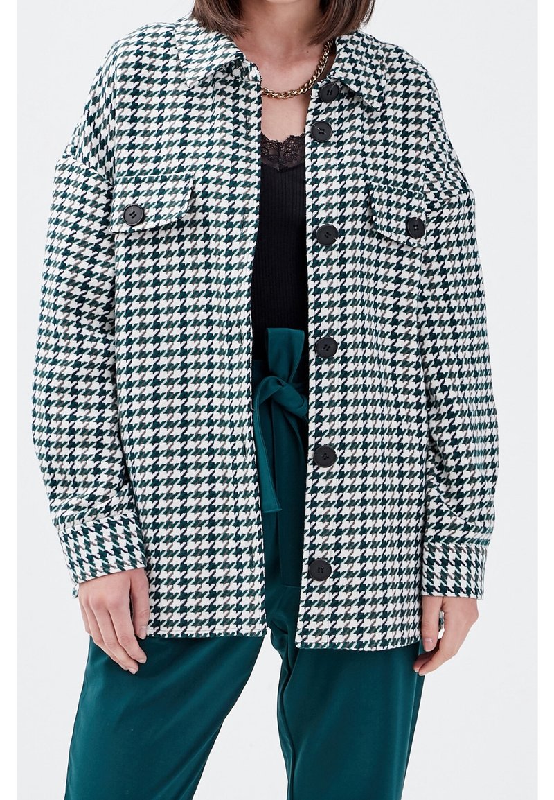 Groen-wit jasje met hondentandpatroon, knoopsluiting, twee voorzakken en een losse pasvorm. Draagt over een zwarte top en turquoise broek.