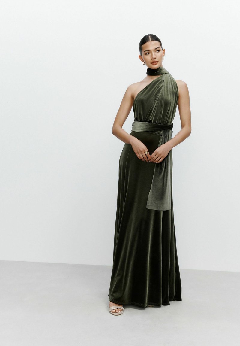 Frau, die ein ärmelloses, dunkelgrünes Samtkleid mit einem trägerlosen, drapierten Design und einem passenden Stoffgürtel trägt, stehend vor einem schlichten Hintergrund.
