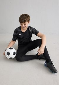 Chlapec v černém sportovním oblečení sedí na podlaze, drží černo-bílý fotbalový míč, usmívá se a dívá se dolů.