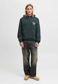 Haut à capuche vert foncé avec une poche avant et du texte imprimé blanc. Assorti à un jean gris clair et des bottes de travail beige.