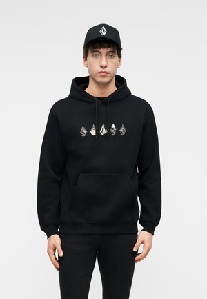 Tânăr purtând un hanorac negru cu modele albe de copaci și geometrice și o șapcă neagră cu un logo geometric alb, stând în picioare pe un fundal simplu.