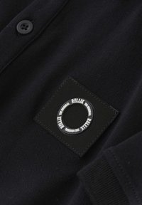 Vêtement en textile noir avec une surface texturée, fermeture à boutons et un patch noir carré avec un accent de logo circulaire en blanc.