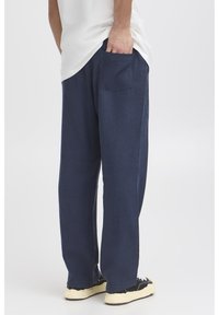 Pantaloni largi, de culoare albastru închis, confecționați dintr-un material texturat, cu un buzunar la spate și tivuri rulate, asortați cu adidași negri.