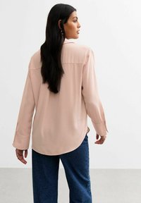 Blush roze blouse met lange mouwen, een losse pasvorm, afgeronde onderrand en knoopsluiting bij de mouwen, gecombineerd met donkerblauwe wijde broek.