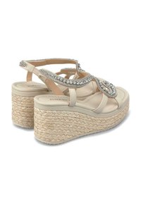 Sandales plateformes beiges avec une base en paille tressée, présentant un design de bride ornée de bijoux et un orteil ouvert. Matériau supérieur lisse et brides de cheville sécurisées.