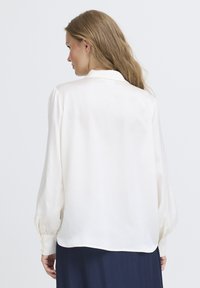 Camicia di seta bianca con una texture liscia, maniche lunghe e polsini con bottoni. Presenta un colletto classico e un design rilassato e fluido.