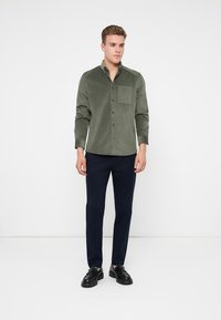 Cămașă verde cu mânecă lungă, dintr-un material texturat, având o buzunar pe piept, asortată cu pantaloni negri închis și pantofi negri.