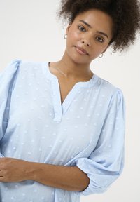 Blouse bleu clair avec des manches bouffantes, tissu texturé présentant de petits points en relief, et un col à encolure décroisée.