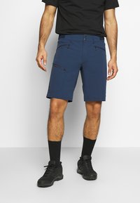 Pantalones cortos azul marino de tejido elástico, con un bolsillo con cremallera en el muslo, cintura plana y un corte clásico. Zapatos y calcetines negros.