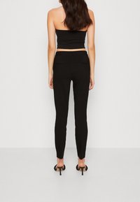 Černé, přiléhavé kalhoty s hladkou texturou a minimalistickým designem, obsahující dvě zadní kapsy a zúžené nohavice, doplněné černými podpatky.