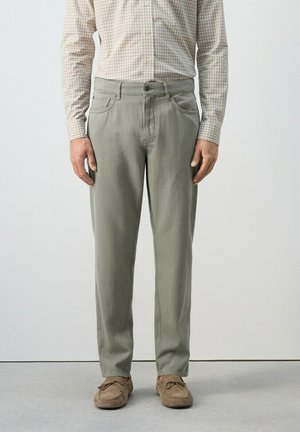 Uomo che indossa pantaloni grigi chiaro a gamba dritta, camicia beige a quadri infilata e mocassini di camoscio marrone, in piedi all'interno.