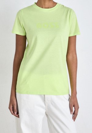 Person trägt ein hellgrünes, figurbetontes kurzärmeliges T-Shirt mit dezentem "BOSS"-Schriftzug und weiße Hose, gezeigt vom Hals bis zur Hüfte.