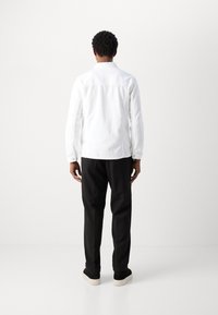 Camicia bianca a maniche lunghe vista da dietro, abbinata a pantaloni neri e calzature nere. Tessuto liscio e uniforme con colletto classico.