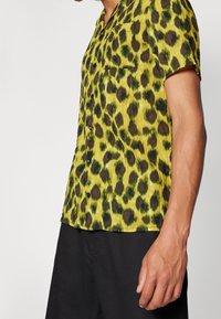 Uomo che indossa una camicia a maniche corte con bottoni, con una stampa leopardata gialla e nera, abbinata a pantaloni neri.