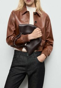 Chaqueta de cuero marrón corta con cuello, botones negros y puños acanalados, combinada con jeans de mezclilla oscura y un bolso marrón oscuro.