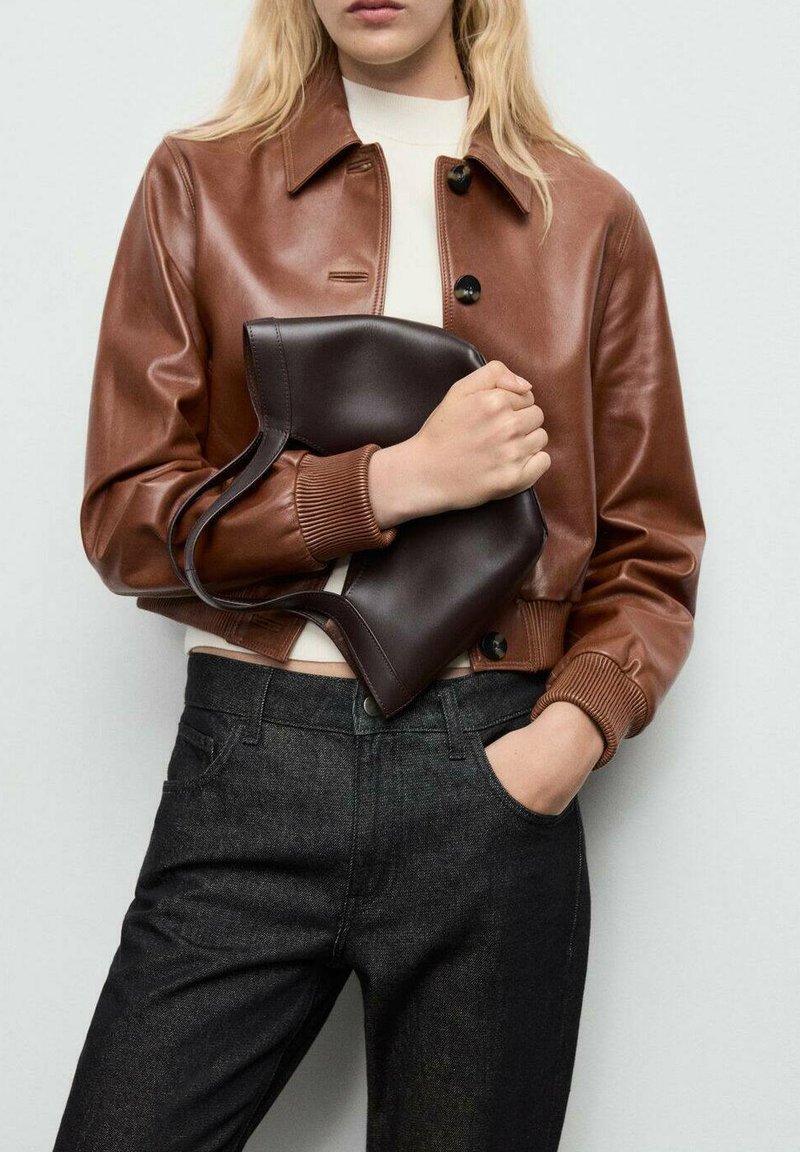 Chaqueta de cuero marrón corta con cuello, botones negros y puños acanalados, combinada con jeans de mezclilla oscura y un bolso marrón oscuro.