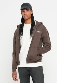 Mladý muž s dlhými vrkočmi, ktorý nosí čiernu čiapku, bielu košeľu, hnedú mikinu McLaren na zips a tmavé džínsy, stojí na jednotnom pozadí.