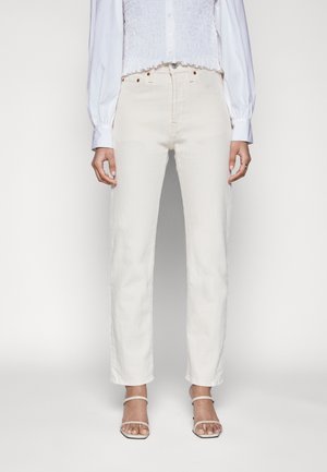 Femme portant un jean blanc droit taille haute, des talons blancs à brides, et une chemise blanche à manches longues boutonnée avec un détail froncé.