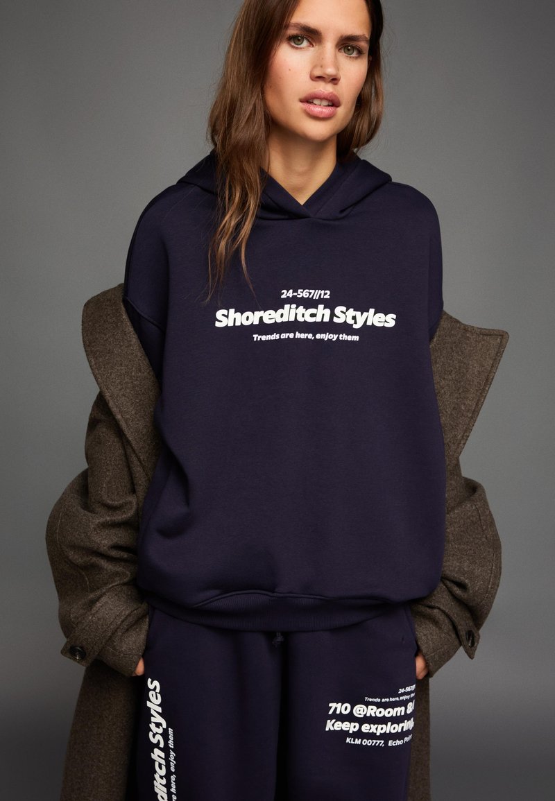 Navy hoodie and matching sweatpants with white text prints. Hoodie features a drawstring and ribbed cuffs. Soft fabric with a relaxed fit.